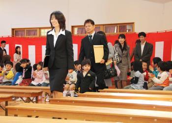 会場内は紅白の幕が掛けられ、長椅子に園児が手を叩きながら笑顔で座っており、新入園児と保護者が並んで入場している入所式の写真