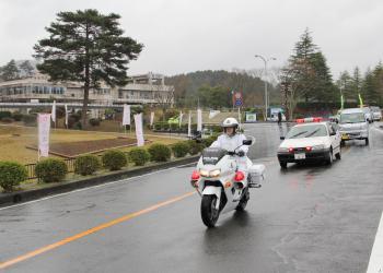 公道を白バイ、その後ろに警察車両が続いて走っている写真
