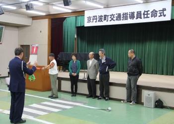 「京丹波町交通指導員任命式」と書かれた横断幕の前に、疑似横断歩道が作られ、警察署員の男性が赤色の信号機を持って立っており、交通指導員の方が横断歩道の前に立って誘導の仕方の指導を受けている様子の写真
