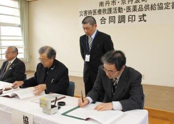 佐々木南丹市長、佐野船井医師会長、寺尾町長が並んで長机に着席しており、寺尾町長が協定書に調印している写真
