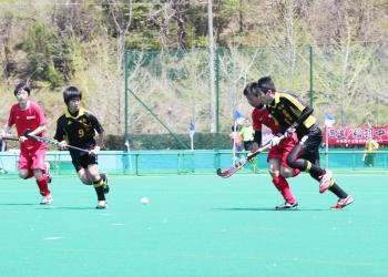 赤いユニフォームを着た選手と黒いユニフォームを着た選手がスティックを持ち、白いボールを奪おうと競い合い走っているホッケーの試合の写真