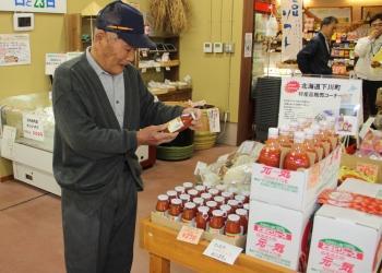 店内の机に、瓶に入ったトマトジュースが沢山陳列されており、帽子を被った年配の男性がトマトジュースを手にとって見ている写真