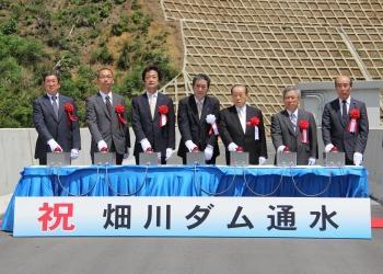 「祝 畑川ダム通水」と書かれた横断幕の後ろの机にボタンが7つ設置され、胸に赤い胸章をつけた6名の関係者の方々と白い胸章をつけた方寺尾町長がボタンに白い手袋をした手をのせている畑川ダム通水式の写真