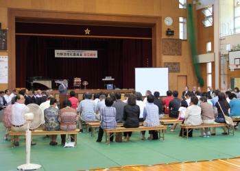 大きな舞台のある体育館のような場所の会場で、舞台の前に長椅子が設置され、来場者が席に着き、設立総会に参加している様子の写真
