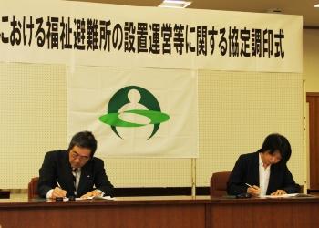 福祉避難所設置運営等に関する協定調印式の横断幕と町章が掲げられているボードの前に寺尾町長と櫻井理事長が席につき、協定書に調印している写真