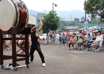 太鼓台に乗せられた大きな和太鼓を、黒い腹掛けの衣装を身に着けた男性がバチを持って全身で叩いて演奏している写真