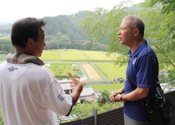 田んぼを見渡せる高所で、田渕代表と祖母井ゼネラルマネージャーが向かい合って話をしている写真