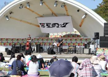ドーム型の野外ステージでギターやドラムを使ったネクターズの演奏が行われており、芝生にシートを広げ、座って音楽を楽しんでいる来場者の皆さんの写真