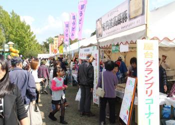 のぼり旗が立てられ、色々な屋台のテントが立ち並んでおり、多くのお客さんで賑わっている会場の様子の写真