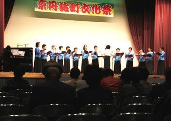 「京丹波文化祭」と書かれた横断幕のある舞台上で、ロングスカートの同じ衣装を着たコーラス・カナリアの皆さんが楽譜を持って、歌を歌っているステージの写真