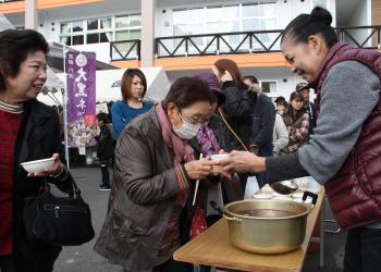 台の上の大きな鍋いっぱいに入っているぜんざい、ふるまいに並んでいる来場者、えんじ色のベストを着た女性が来場者のマスクを付けた女性に振る舞いのぜんざいを手渡している写真