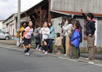 肩にタスキを掛けて沿道を走っているランナーと沿道に立って手を叩いたり、声を掛けて声援を送っている人々の写真