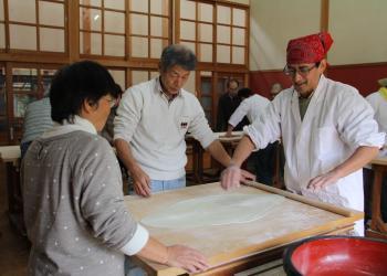 四角の麺打ち台に下打ち粉が薄くふられ、赤い三角巾を頭に巻いた男性が、のし棒を使って、薄くそばの生地を伸ばしている写真