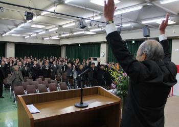 舞台上に立つ吉川文則区長会長が両手を上に挙げて万歳三唱をしており、会場内にいる多くの出席者も会長に合わせて両手を挙げて万歳三唱をしている写真