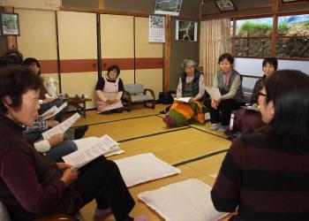和室の会場で、参加者が輪になって座っており、資料を手に持ち保健師の女性の話を興味深く聞いている写真