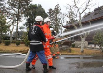 ヘルメットを被り、半纏を着た男性、ヘルメットを被り、オレンジ色の防火服を着た男性が、消防ホースを持ち、水が勢いよく放水されている防火訓練の写真