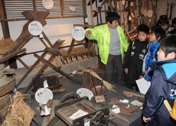 昔の生活の道具が沢山展示されており、黄色いジャンパーを着た運営委員の女性が見学に訪れた児童達に説明をしている写真