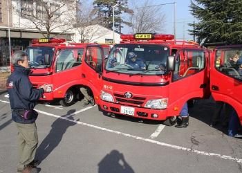 駐車場に停まっている赤い消防車両、車両前に立って話をしている納入業者の男性、消防車両の運転席の扉が開き、団員が運転席の車内を確認している写真