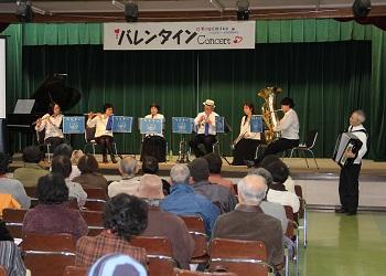 「バレンタインConcert」と書かれた横断幕のある舞台上で、フルートやトランペット、アコーディオンなどの楽器を演奏しており、沢山の来場者が席に着いて演奏を聞いているバレンタインコンサートの写真