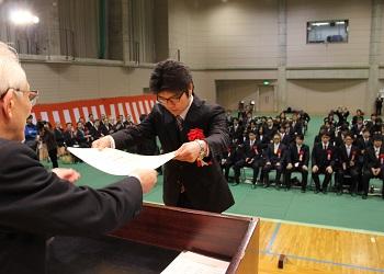 卒業生、参列者が会場内で椅子に着席しており、胸に赤い花の胸章を付けた卒業生の男性が、舞台上の演台の前で、卒業証書を両手で受け取っている写真