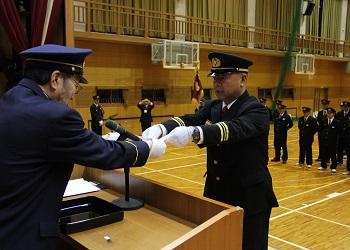 消防の帽子と制服を着た梅原団長が舞台上の演台の前で、寺尾町長から任命書を受けとっている写真