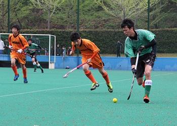 緑色のユニフォームを着た選手がスティックで黄色いボールをコントロールしながら走っており、オレンジ色のユニフォームを着てスティックを持った選手が、黄色いボールを狙って追いかけているホッケーの試合の写真