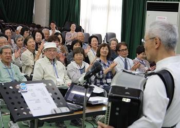 受講者の前で米村さんがアコーディオンを肩にかけて演奏しており、受講者の皆さんが手拍子をしながら歌を歌っている写真