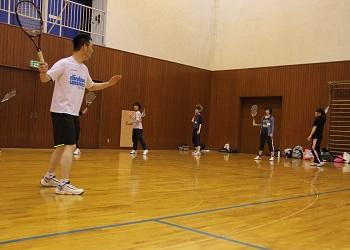 体育館で、テニスのラケットを持った受講者が間隔をあけて広がっており、白いTシャツを着た講師の男性から指導を受けているテニス教室の写真