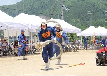 ヘルメットをかぶりオレンジ色のゼッケンをつけ、紺色の活動服を着た男性2名が素早い動きで、消防用の筒状の吸管の管を持ち上げて伸ばす操法技術を披露している写真