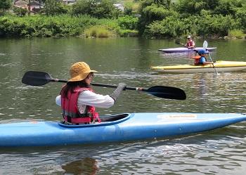 青いカヌーに乗り両手でパドルをもって水面を漕いでいる女性と川の向こうにも2艘のカヌーに乗っている体験者が写っている写真