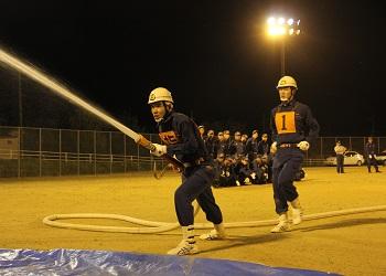 ヘルメットをかぶり活動服を着た男性が足を前後に大きく開き腰を落として消防ホースを持ち、ヘルメットをかぶり活動服を着て黄色いゼッケンをつけた男性がすぐ後ろに駆け寄ってきており、ホースから勢いよく水が放水されている写真
