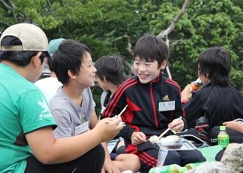 児童達が、レジャーシートに座り、笑顔で楽しそうに会話を楽しみながらお弁当を食べている写真