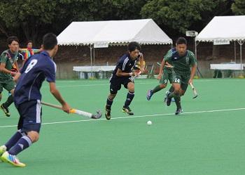 紺色のユニフォームを着た選手と緑色のユニフォームを着た選手がスティックを左手に持ち、前方にある白いボールを奪うために競い、走っているホッケーの試合の写真