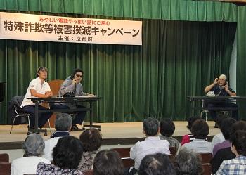 「特殊詐欺被害撲滅キャンペーン」と書かれた横断幕、舞台左側の長机に座るにサングラスをかけ電話をしている男性と白いポロシャツの男性、右側の長机に座り、電話をしている黄色いバンダナを頭に巻いた男性が、寸劇で詐欺の手口を紹介しており、たくさんの来場者が会場の席について寸劇を見ている写真