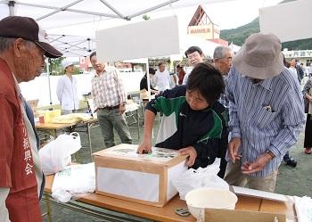 中央に丸い穴の開いた長方形の箱が机の上に置かれ、ジャージ姿の来場者が穴の中に手を入れて栗のつかみ取りに挑戦しており、後ろに立つ男性がつかみ取りの様子を覗き込んで見ている写真