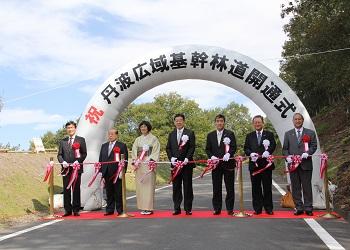 「祝 丹波広域基幹林道開通式」と書かれた半円型 エアーアーチ門の前で市長と6名の関係者がテープカットをしている写真