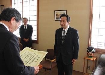 株式会社京都環境保全公社の檀野恭介社長と寺尾町長が向かい合って立っており、寺尾町長が賞状を読み上げて表彰している写真