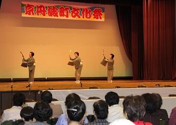 「京丹波文化祭」と書かれた横断幕のある舞台上で、着物を着た3人の女性が、扇子を右手に持って上にあげ、左手を肘に添えて踊りを披露している写真