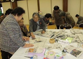 高齢の女性が集まっており、机の上にたくさんの新聞紙がおかれ、中央に立つ女性が他の来場者に新聞紙を指差しながら作り方の説明をしている写真