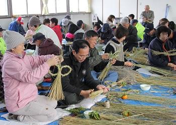 会場いっぱいに参加者が集まり、ビニールシートに座って藁を束ねたり、ねじったりしてしめ縄づくりの作業をしている写真
