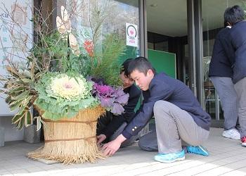おおきなハボタン、松、竹、南天などの材料で作られた立派な門松を須知高校生の生徒が玄関前に設置している写真
