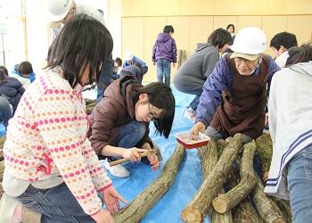 指導者の年配の男性に見守られながら、女子児童が真剣な表情で金づちを使って、原木に種菌を打ち込んでおり、その様子を他の児童がみている写真