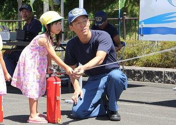 消防員の男性と一緒に女の子が赤い消火器のノズルを持っており、赤い消火器のノズルから消火剤が噴出している初期消火の体験をしている写真