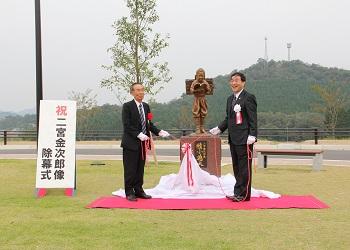 「祝二宮金次郎像除幕式」と書かれた看板の横に柴を背中に背負い、本を読んでいる二宮金次郎像が設置されており、片山山治さんと町長が白い布についている赤いリボンを持って除幕式を行っている写真