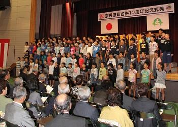 「祝 京丹波町合併10周年記念式典」と書かれた横断幕の前に町内の4年生が整列して合唱を披露しており、来場者が椅子に座って歌を聞いている式典の様子の写真