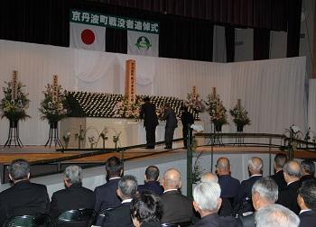 舞台の壁に白い布が掛けられ、きれいに飾られた祭壇、追悼の碑が舞台中央に設置され、3名の方が献花をしている写真