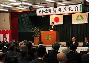「京丹波町新春互礼会」と書かれた横断幕、日の丸の国旗、京丹波町章が舞台上に掲げられ、演台に立ってあいさつを述べている町長、町内各団体の代表が席について話を聞いている様子を後方から写した写真