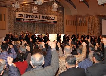 「2016北陸新幹線小浜ルート建設促進総決起大会」と書かれた横断幕が舞台上に掲げられ、舞台に立つ多くの関係者と沢山の参加者が右手の握りこぶしを上にあげて、「ガンバロー」を三唱している写真