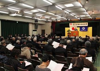 「祝 設立10周年記念式典」と書かれた横断幕、金屏風が舞台に設置され、講演台に立って話をしている男性、多くの会員の方々が席につい資料に目を通しながら話を聞いている式典の様子の写真
