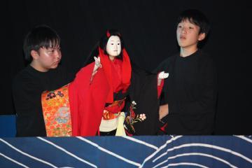 2人の男子生徒が赤い着物を着た人形を操って演じている、人形芝居の舞台の写真